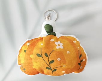 Puede incluir: Un llavero de calabaza de peluche con cuerpo naranja, detalles florales blancos y tallo verde. La calabaza tiene un llavero plateado adjunto en la parte superior. El diseño incluye un estampado floral con pequeñas flores blancas y hojas verdes.