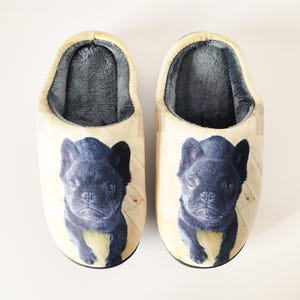 May include: A pair of cozy slippers featuring a black French bulldog print. The slippers have a light-colored exterior with a wooden floor pattern and a soft, gray interior. These house shoes are designed for comfort and style.