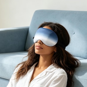 May include: A sleep mask featuring an ombre gradient pattern that transitions from a deep navy blue at the top to a soft white at the bottom. The mask has a smooth, contoured shape designed to cover the eyes and is secured with a black elastic strap.