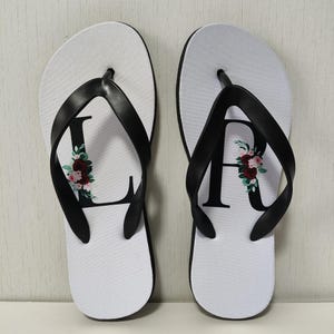 May include: A pair of white flip-flops with black straps. Each flip-flop features a large black letter, adorned with a floral design in shades of red, pink, and green. The flip-flops are designed for casual wear.