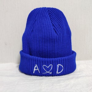 Puede incluir: Un gorro de punto azul con un corazón bordado en blanco y las letras "A♥D" en el puño.