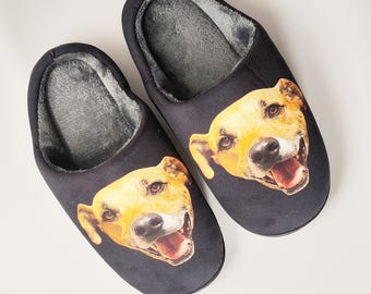 May include: A pair of black slip-on slippers featuring a printed graphic of a tan dog's face on the top of each shoe. The interior of the slippers is lined with a soft, grey plush material, providing a contrast to the dark exterior fabric.