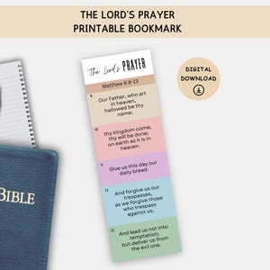 May include: A printable bookmark featuring "The Lord's Prayer" from Matthew 6:9-13, with a colorful, segmented design. The text is displayed in a clear, readable font. A spiral notebook, a pencil, and a blue Holy Bible are also visible.