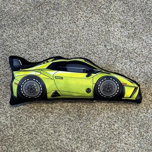 May include: A green and black car-shaped pillow. The pillow is a side view of a sports car with black wheels and a black interior.