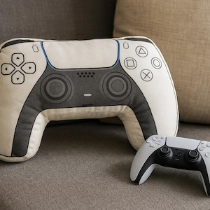 May include: A large, cream-colored controller-shaped pillow with black accents and button details, alongside a smaller, matching controller. The design mimics a gaming controller, perfect for gamers. The items are on a gray couch.