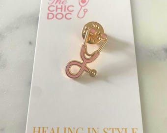 May include: A gold-colored stethoscope pin with pink enamel detailing, resting on a white card. The card features the text "The Chic Doc" and "Healing in Style" in gold lettering. The pin is a medical-themed accessory.