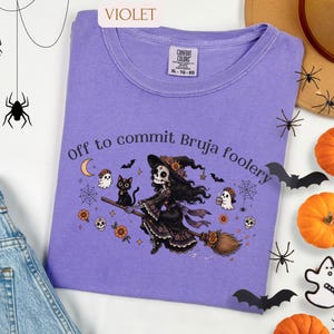 May include: A violet t-shirt featuring a graphic of a skeleton witch flying on a broomstick with a black cat, ghosts, and flowers. The text reads "Off to commit Bruja foolery". The shirt has a crew neckline and a Comfort Colors brand label.