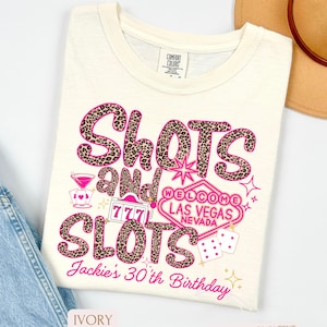 May include: Ivory t-shirt with a pink and black leopard print design that says "Shots and Slots" with a Las Vegas casino sign and dice. The text "Jackie's 30th Birthday" is at the bottom of the shirt.