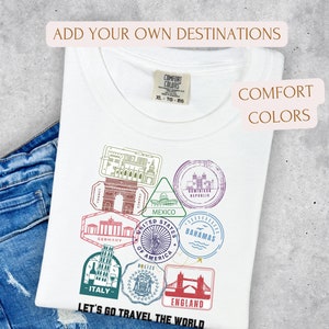 May include: White t-shirt with a travel-themed graphic featuring stamps from various countries, including France, Germany, and Italy. The shirt also has the text "Let's Go Travel The World" and "Add Your Own Destinations".