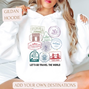 May include: White Gildan hoodie featuring travel-themed stamps with destinations like Italy, England, and Mexico. The phrase "Let's Go Travel The World" is printed below the stamps. The hoodie also includes the text "Add Your Own Destinations".