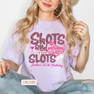 May include: Purple t-shirt with a leopard print design that says "Shots and Slots" with a Las Vegas sign and dice. The text "Jackie's 30th Birthday" is at the bottom of the shirt.