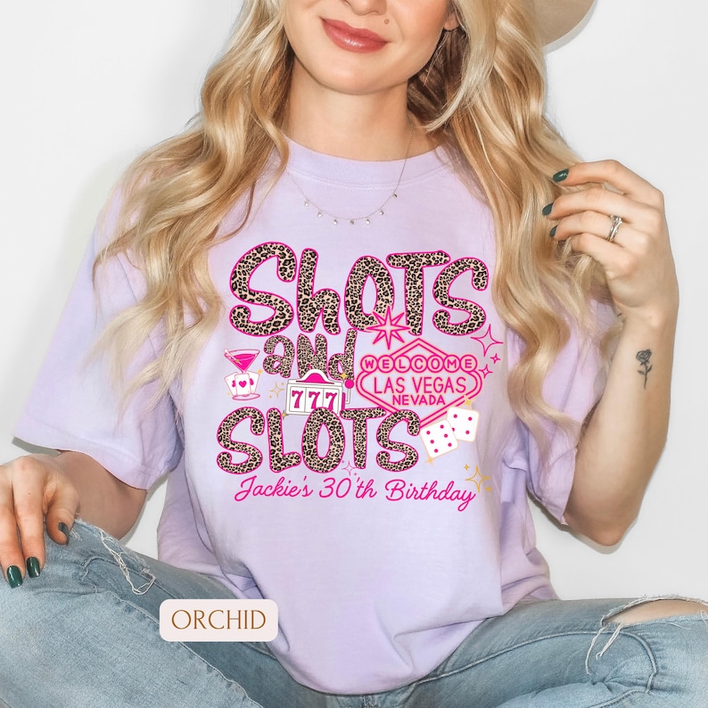 May include: Purple t-shirt with a leopard print design that says "Shots and Slots" with a Las Vegas sign and dice. The text "Jackie's 30th Birthday" is at the bottom of the shirt.