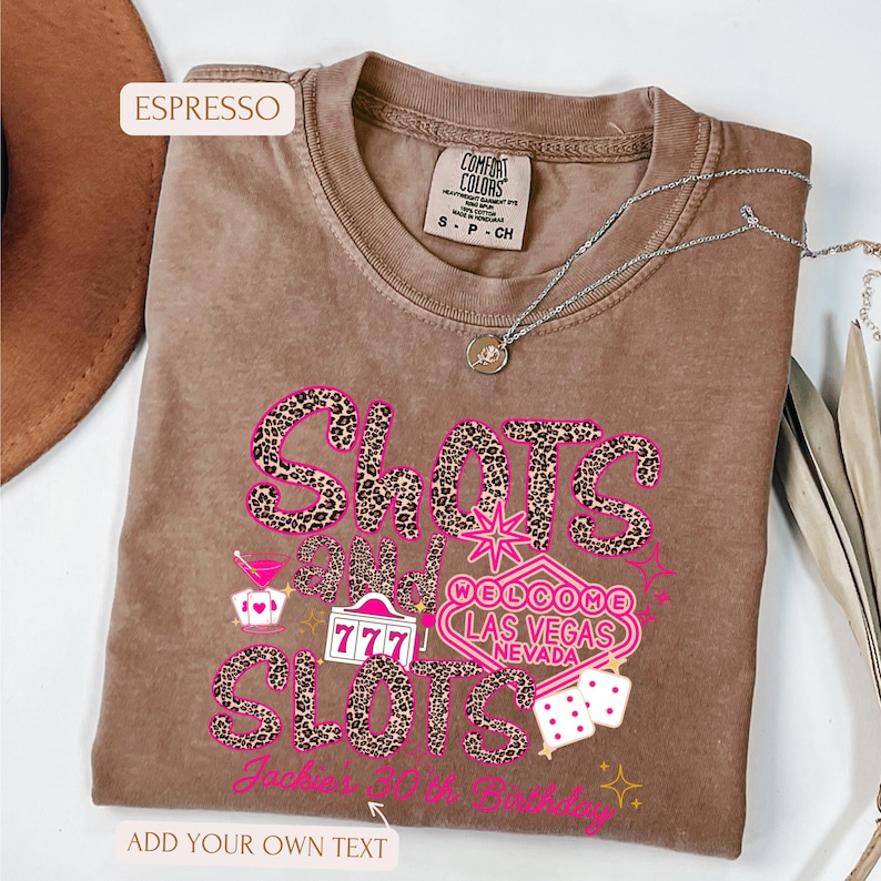 May include: A brown t-shirt with a pink and black leopard print design that says "Shots and Slots" with a Las Vegas welcome sign and dice. The text "Jack's 30th Birthday" is at the bottom of the shirt.