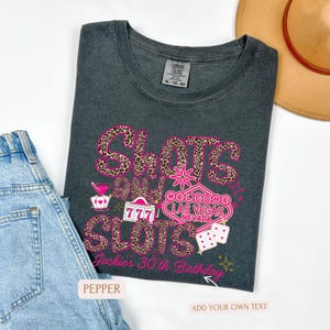 May include: A dark gray t-shirt with a pink and leopard print design that says "Shots and Slots" with a Las Vegas sign that says "Welcome to Las Vegas, Nevada". The shirt also has a pink and black dice graphic and the text "Jackie's 30th Birthday".