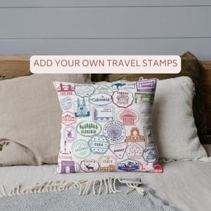 May include: A white square pillow with colorful travel stamp designs from various countries. The pillow features stamps from England, Mexico, Colombia, and more. Text on the pillow reads "ADD YOUR OWN TRAVEL STAMPS".