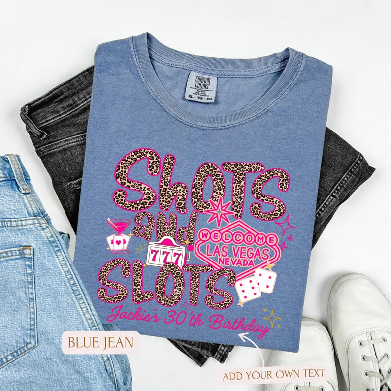 May include: A blue t-shirt with a pink and leopard print design that says "Shots and Slots" with a Las Vegas sign and dice. The text "Jackie's 30th Birthday" is also on the shirt.