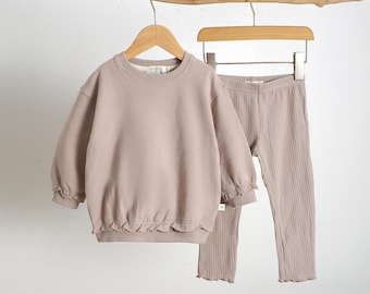 May include: A two-piece children's outfit in a neutral taupe color. The set includes a long-sleeved sweatshirt with a ruffled hem and cuffs, and ribbed leggings. The sweatshirt has the text "almond RUD" on the tag. The outfit is hanging on a wooden branch.
