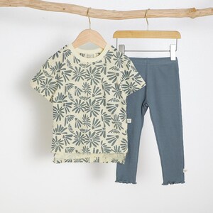 May include: A children's two-piece outfit featuring a short-sleeved t-shirt and leggings. The t-shirt is cream-colored with a gray floral pattern. The leggings are a solid blue-gray color with a ruffled hem. The outfit is hanging on a wooden hanger.