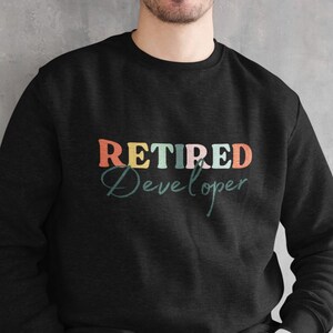 May include: Black sweatshirt with the text "RETIRED Developer" in colorful letters. The word "RETIRED" is in orange, red, and blue, while "Developer" is in a teal script font. The sweatshirt is a crew neck style.