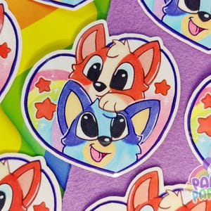May include: A heart-shaped sticker featuring a cartoon illustration of a red dog and a blue dog stacked on top of each other. The background inside the heart includes a colorful rainbow and several red stars. The text Panda Parfait is visible.
