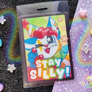 K&ouml;nnte beinhalten: Ein farbenfroher Comic-Charakter mit einer Partym&uuml;tze und einer Clownsnase. Der Charakter h&auml;lt ein Schild mit der Aufschrift "Stay Silly!" Der Hintergrund ist ein gelber und gr&uuml;ner Aquarell-Wasch.