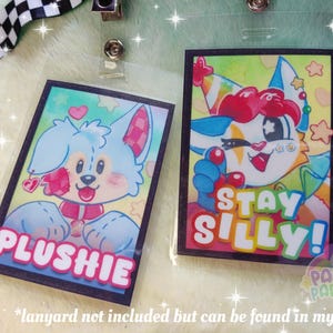 K&ouml;nnte beinhalten: Zwei farbige Cartoon-Charakter-Badgehalter mit schwarzen R&auml;ndern. Der erste Badgehalter zeigt einen blauen und wei&szlig;en Cartoon-Charakter mit rosa Akzenten und dem Text "PLUSHIE". Der zweite Badgehalter zeigt einen roten, wei&szlig;en und gelben Cartoon-Charakter mit dem Text "STAY SILLY!"