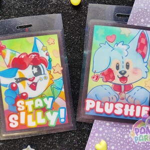 K&ouml;nnte beinhalten: Zwei bunte Badge-Halter mit Cartoon-Charakteren auf wei&szlig;em Hintergrund. Der linke Badge-Halter zeigt einen Cartoon-Charakter mit einem rot-wei&szlig;-blauen Hut und dem Text "Stay Silly!" Der rechte Badge-Halter zeigt einen blauen Cartoon-Charakter mit einer rot-wei&szlig; karierten Schleife und dem Text "Plushie".