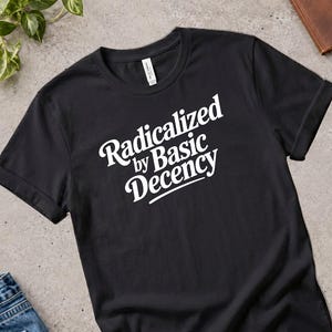 Puede incluir: Una camiseta negra de manga corta con texto blanco impreso en el pecho que dice "Radicalized by Basic Decency" en una fuente negrita y estilizada. La prenda tiene cuello redondo y un corte estándar con un diseño limpio y sencillo.