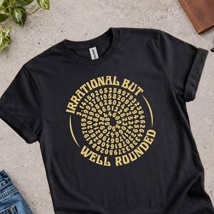 Peut inclure: Un t-shirt noir à manches courtes avec un motif graphique doré. L'impression affiche le texte "IRRATIONAL BUT WELL ROUNDED" entourant une disposition en spirale des chiffres de pi, commençant au centre par le nombre 3.14.