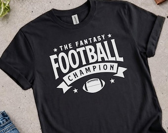 Può includere: Una t-shirt nera a maniche corte con testo stampato in bianco che recita THE FANTASY FOOTBALL CHAMPION. Il design include la grafica di un pallone da football americano centrato sotto il testo, affiancato da piccole stelle su uno sfondo a nastro.