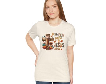 May include: A cream-coloured t-shirt with a graphic design featuring a cross, a pumpkin spice latte, pumpkins, and autumn leaves. The text reads "Pumpkin Spice & Jesus Christ". The shirt is worn by a person.
