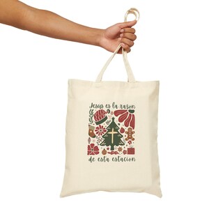 May include: A cream-coloured tote bag featuring a Christmas design. The design includes a Christmas tree, a cross, and the text "Jesus es la razon de esta estacion". The bag is held by a person.