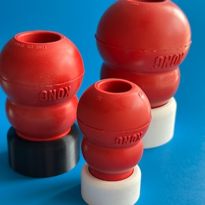 Puede incluir: Tres juguetes para perros Kong de goma roja de diferentes tamaños. El juguete más grande está sobre una base negra, el juguete mediano está sobre una base blanca y el juguete más pequeño está sobre una base blanca. Los juguetes son todos rojos y tienen la palabra "Kong" impresa en ellos.