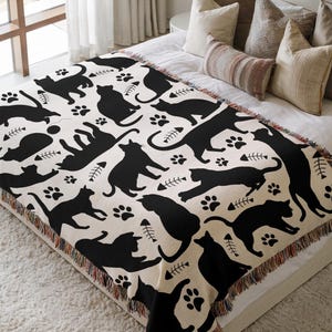 May include: A cream-colored throw blanket featuring a repeating pattern of black cat silhouettes, paw prints, and fish bones. The blanket has a fringed edge and is displayed on a bed with pillows.