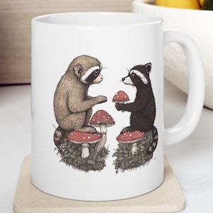 May include: White ceramic mug with a graphic of two animals, a raccoon and a monkey, sitting on mushrooms. The animals are facing each other and appear to be sharing mushrooms.
