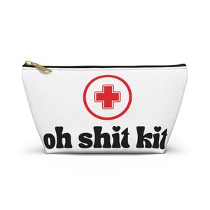 May include: White zippered pouch with a red cross symbol inside a red circle. The words "oh shit kit" are printed in black, with hearts replacing the periods. The pouch has a gold zipper.