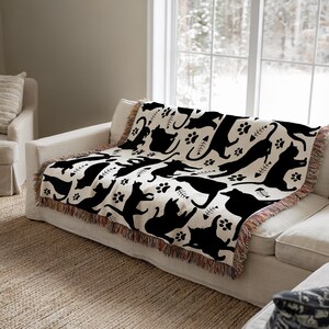 May include: A cream-colored throw blanket featuring a repeating pattern of black cat silhouettes, paw prints, and fish bones. The blanket has a fringed edge and is draped over a white sofa in front of a window.