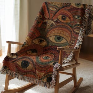May include: A woven throw blanket draped over a wooden rocking chair. The blanket features a repeating pattern of stylized eyes in shades of brown, orange, and green. The blanket has a fringed edge.