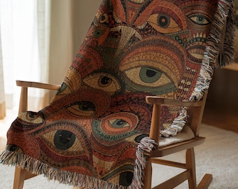 May include: A woven throw blanket featuring a repeating pattern of stylized eyes in shades of orange, brown, yellow, and green. The blanket has a textured surface with intricate line work and decorative fringe along the edges, draped over a wooden chair.