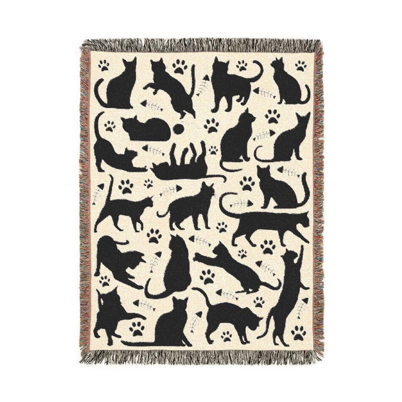 May include: Cream-colored throw blanket with a repeating pattern of black cat silhouettes, paw prints, and fish bones. The blanket has a fringed edge and is suitable for a bed or sofa. The cats are in various poses.