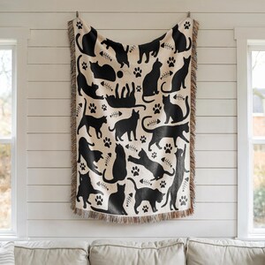May include: A cream-colored throw blanket featuring a repeating pattern of black cat silhouettes, paw prints, and fish bones. The blanket has a fringed edge and is hanging on a wall. The design includes various cat poses.