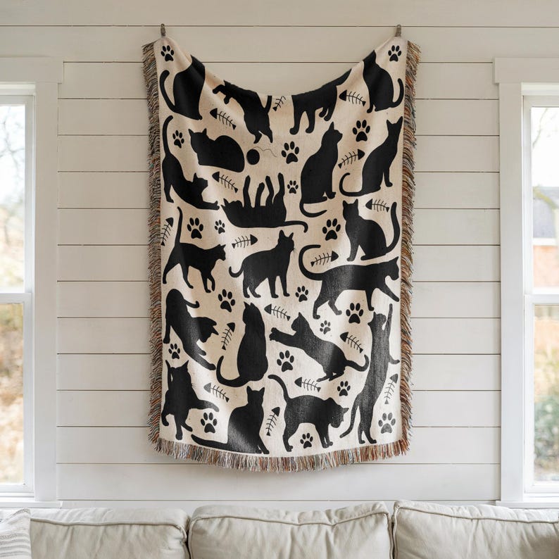 May include: A cream-colored throw blanket featuring a repeating pattern of black cat silhouettes, paw prints, and fish bones. The blanket has a fringed edge and is hanging on a wall. The design includes various cat poses.