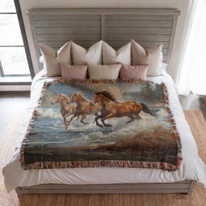 May include: A woven throw blanket featuring a painting of three brown horses running through water. The blanket has a fringed edge and is displayed on a bed with pillows.