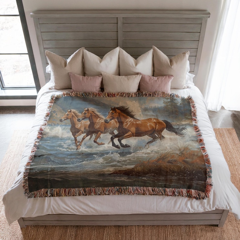 May include: A woven throw blanket featuring a painting of three brown horses running through water. The blanket has a fringed edge and is displayed on a bed with pillows.
