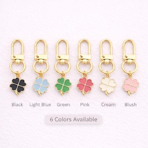 May include: Six gold-toned keychains with four-leaf clover charms in various colors: black, light blue, green, pink, cream, and blush. Each charm is attached to a gold clasp. The text "6 Colors Available" is below the keychains.