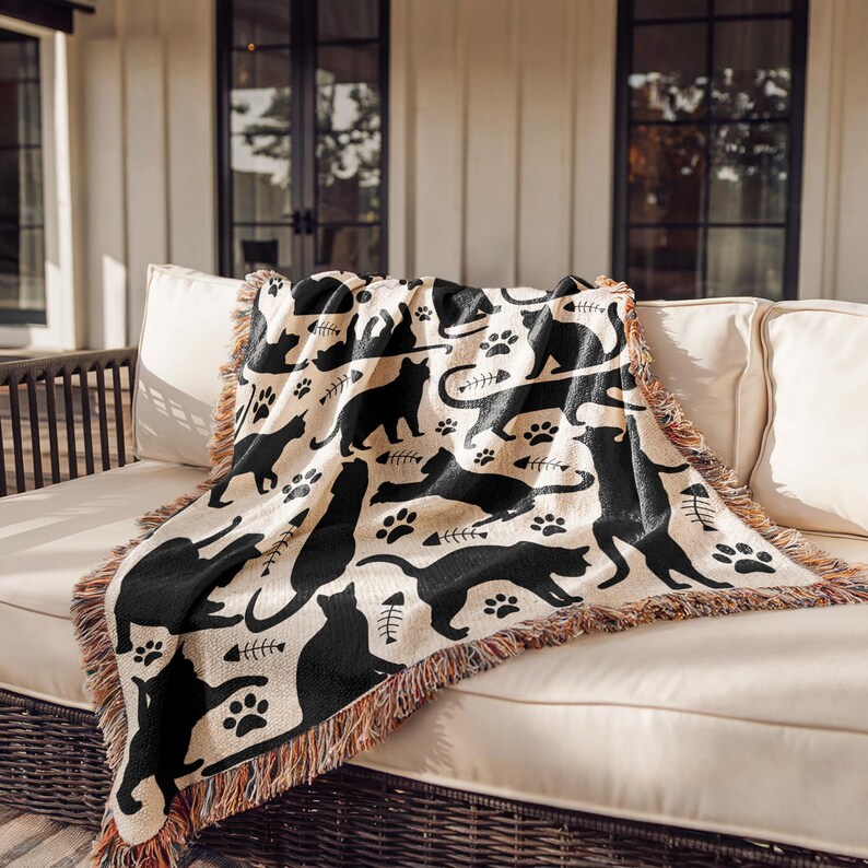 May include: A beige throw blanket with a repeating pattern of black cat silhouettes, paw prints, and fish bones. The blanket has a fringed edge in shades of brown and tan. The blanket is draped over a white couch.