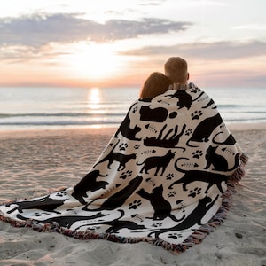 May include: A black and white cat-themed blanket with fringe, featuring silhouettes of cats, paw prints, and fish bones. The blanket is draped over two people on a sandy beach, with the ocean and sunset in the background.