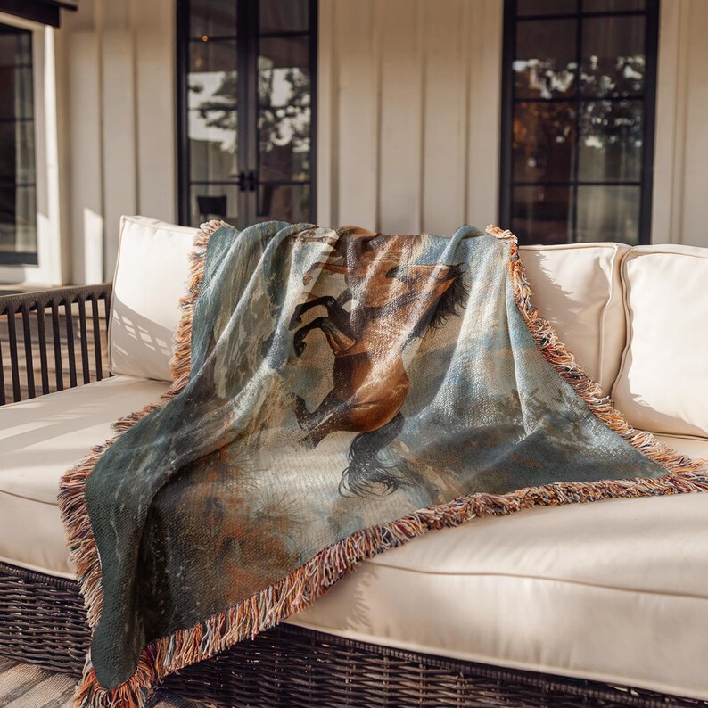 May include: A woven throw blanket featuring a horse illustration in shades of brown, blue, and green. The blanket has a fringed edge in brown and tan and is draped over a beige outdoor sofa. The throw is a decorative home accent.
