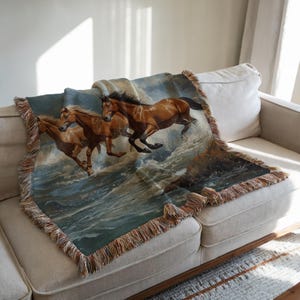 May include: A woven throw blanket featuring a detailed image of three brown horses galloping through ocean waves. The blanket has a fringed edge in shades of brown and tan. The horses are depicted with flowing manes and tails, set against a backdrop of blue and white water.