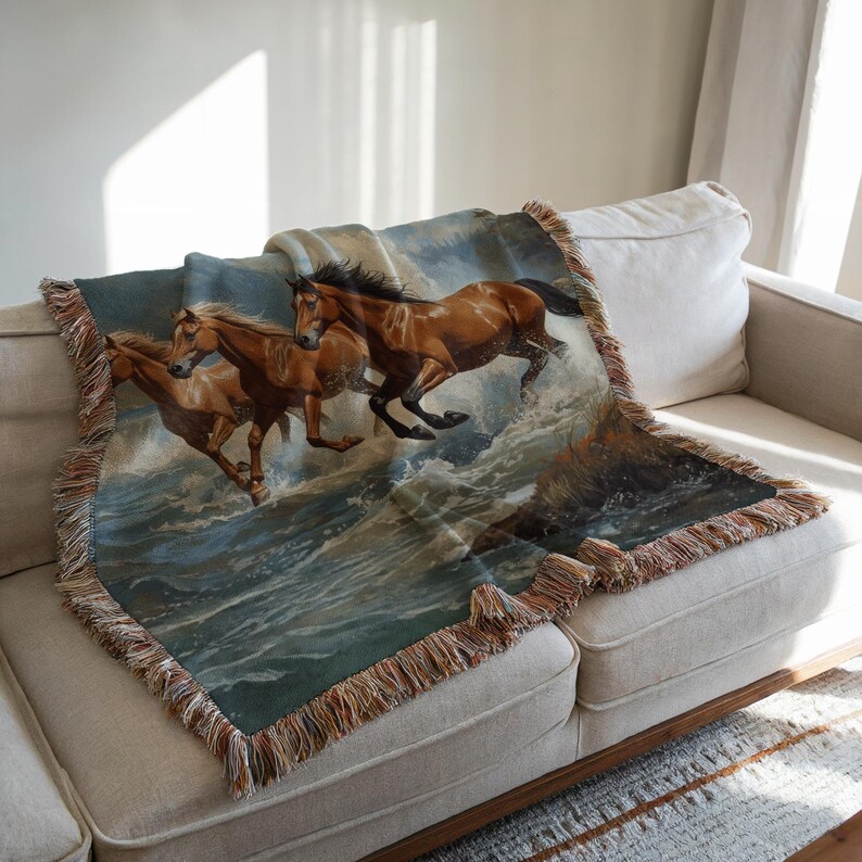 May include: A woven throw blanket featuring a detailed image of three brown horses galloping through ocean waves. The blanket has a fringed edge in shades of brown and tan. The horses are depicted with flowing manes and tails, set against a backdrop of blue and white water.
